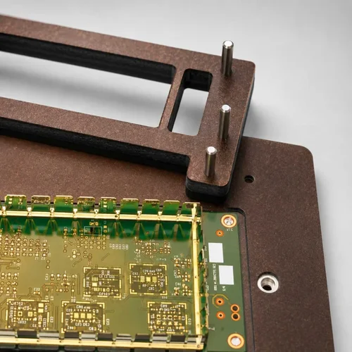 Close-up of a brown composite SMT carrier holding a green PCB, with a matching brown holddown frame with three vertical guide pins positioned above it